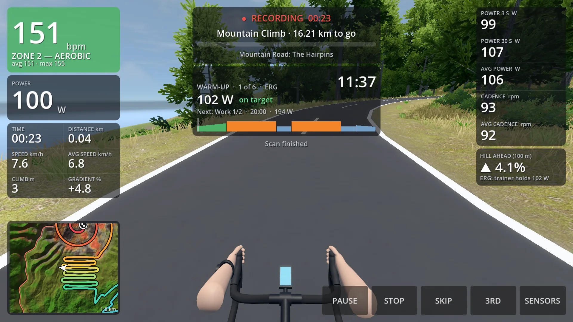 Riding up the island's mountain road in first person: handlebars in view, with heart rate, power, the hill ahead, a workout's steps and a minimap on the screen.
